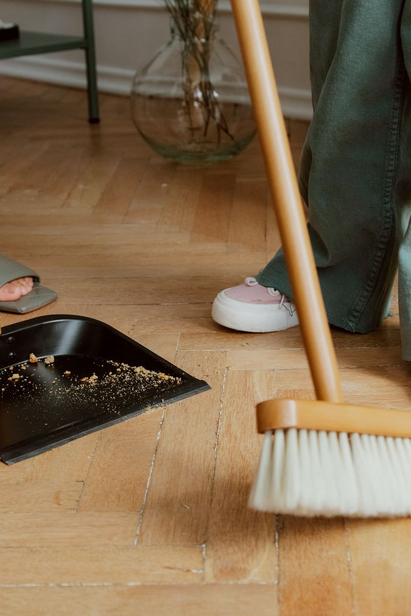 Close-up of people cleaning wooden floor with broom and dustpan indoors.